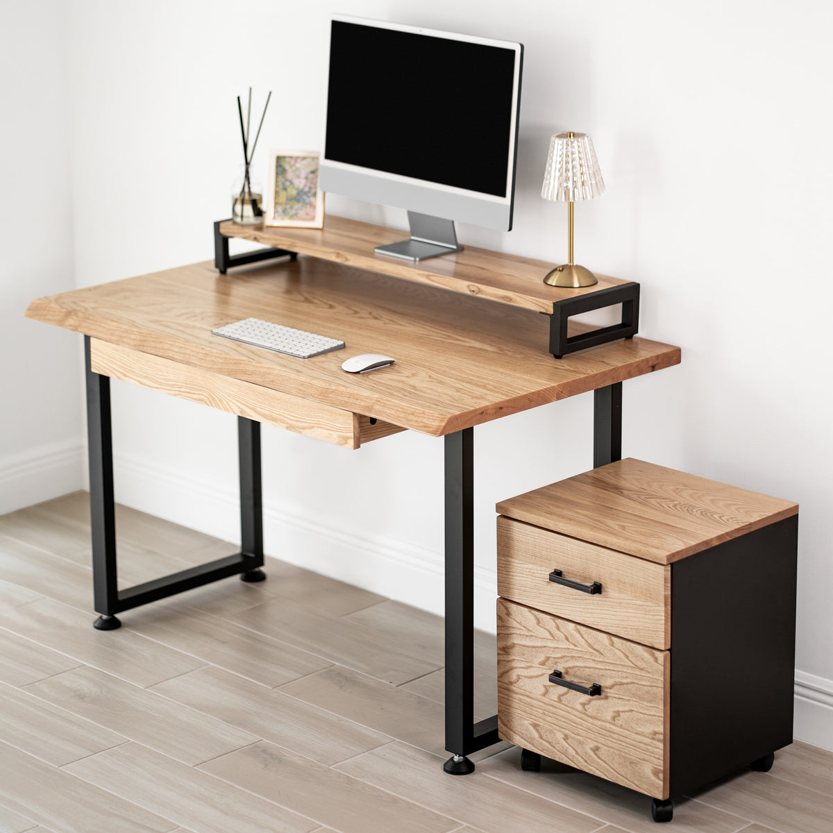 White oak computer desk with a drawer, black metal legs, and a matching storage unit against a white wall.