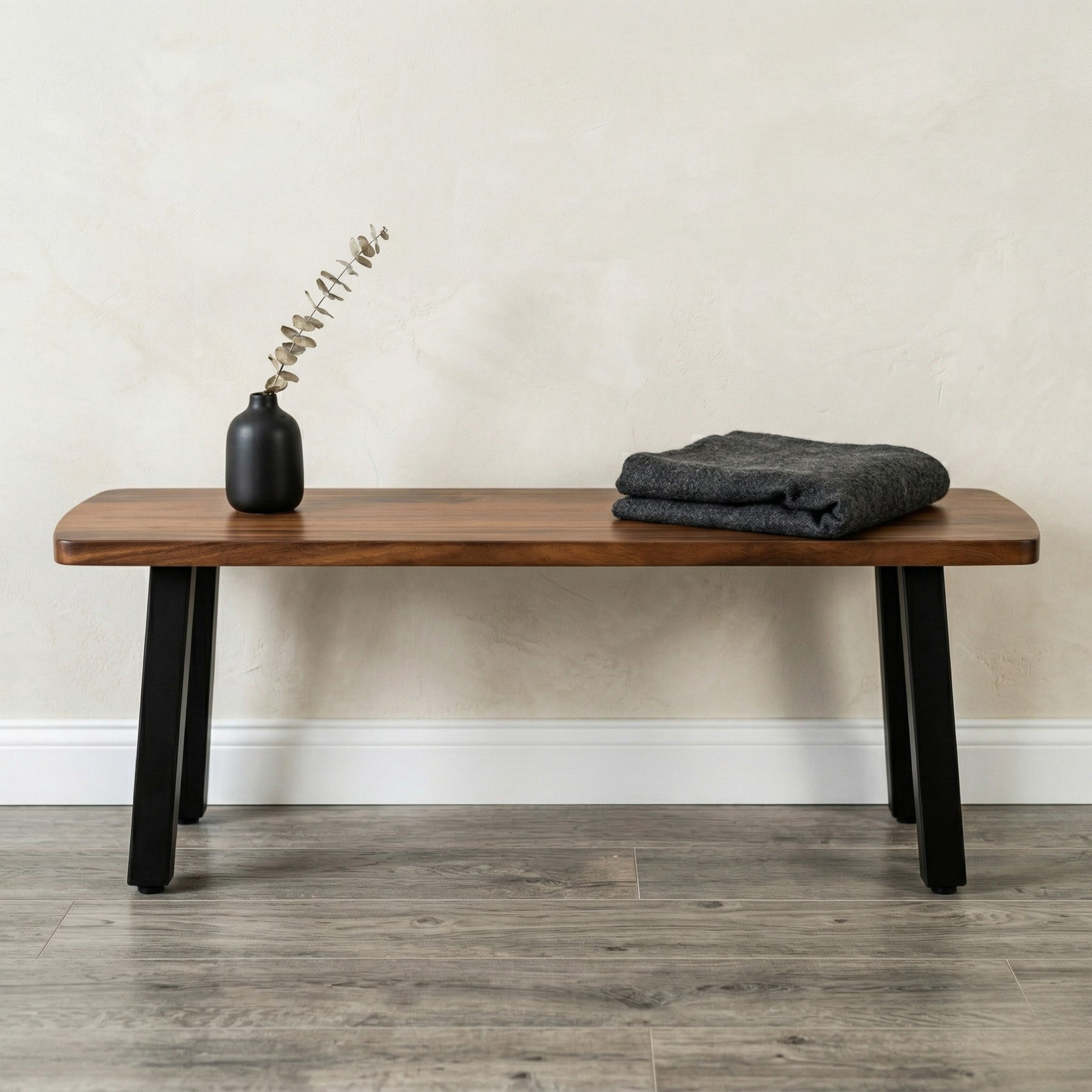 Walnut solid wood bench with black legs, a vase, and folded fabric against a neutral wall.