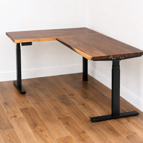 Walnut L-shaped standing desk with black adjustable legs on a wooden floor.