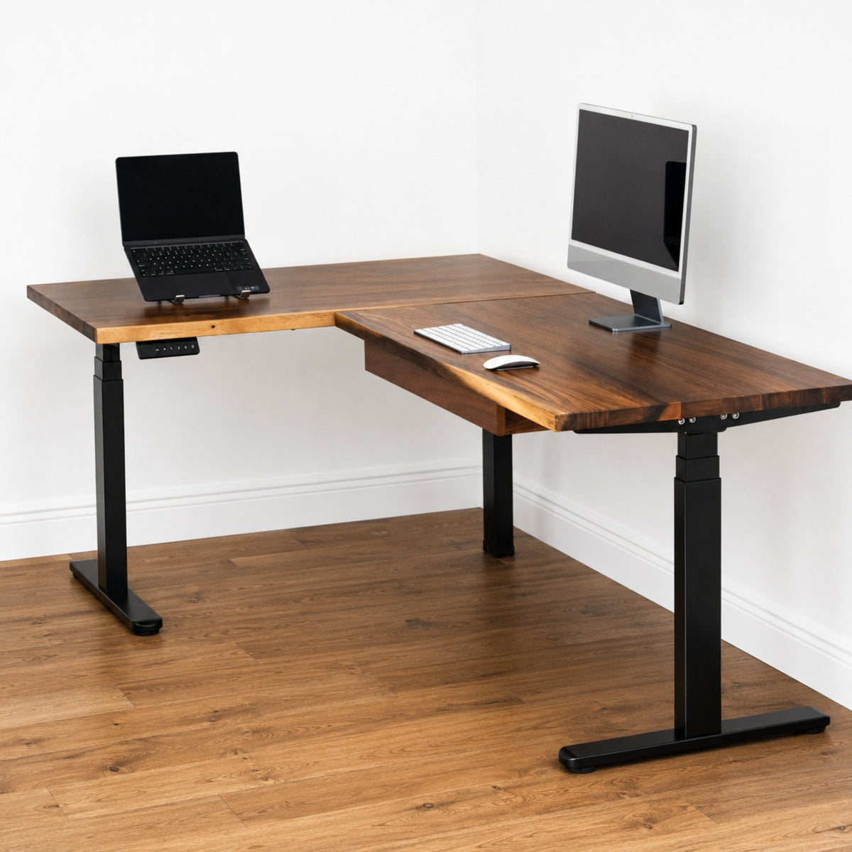 Walnut L-shaped standing desk with a laptop, keyboard, and monitor on a wooden floor.