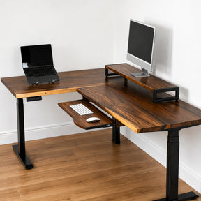 Walnut L-shaped standing desk with laptop, monitor, and keyboard on a wooden floor.