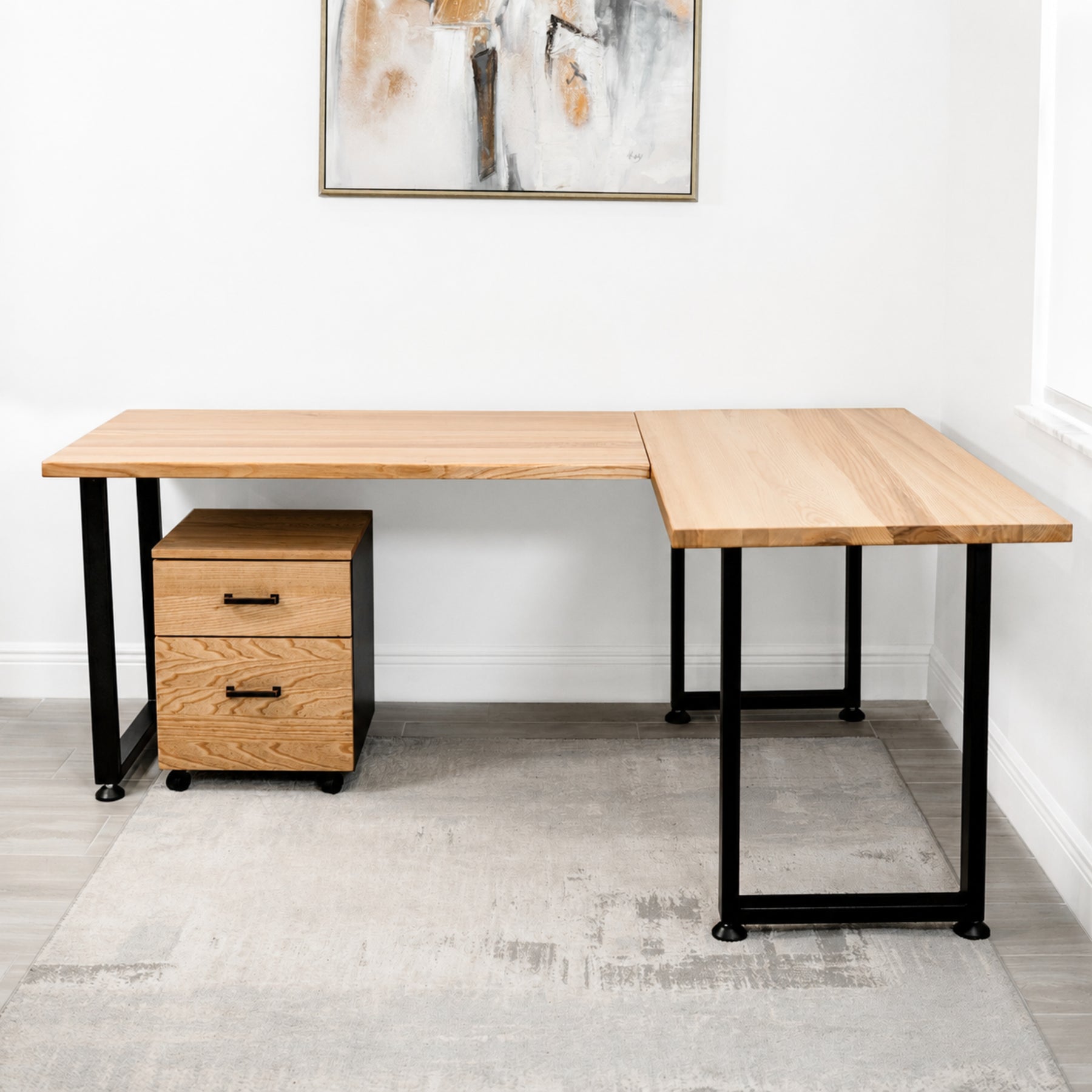 White oak L-shaped desk with black metal legs in a room with abstract art on the wall.