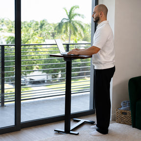 Man working while standing at a height-adjustable walnut laptop desk beside a bright window