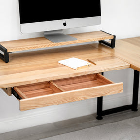 White oak L-shaped desk with a monitor and open drawer on a light gray background