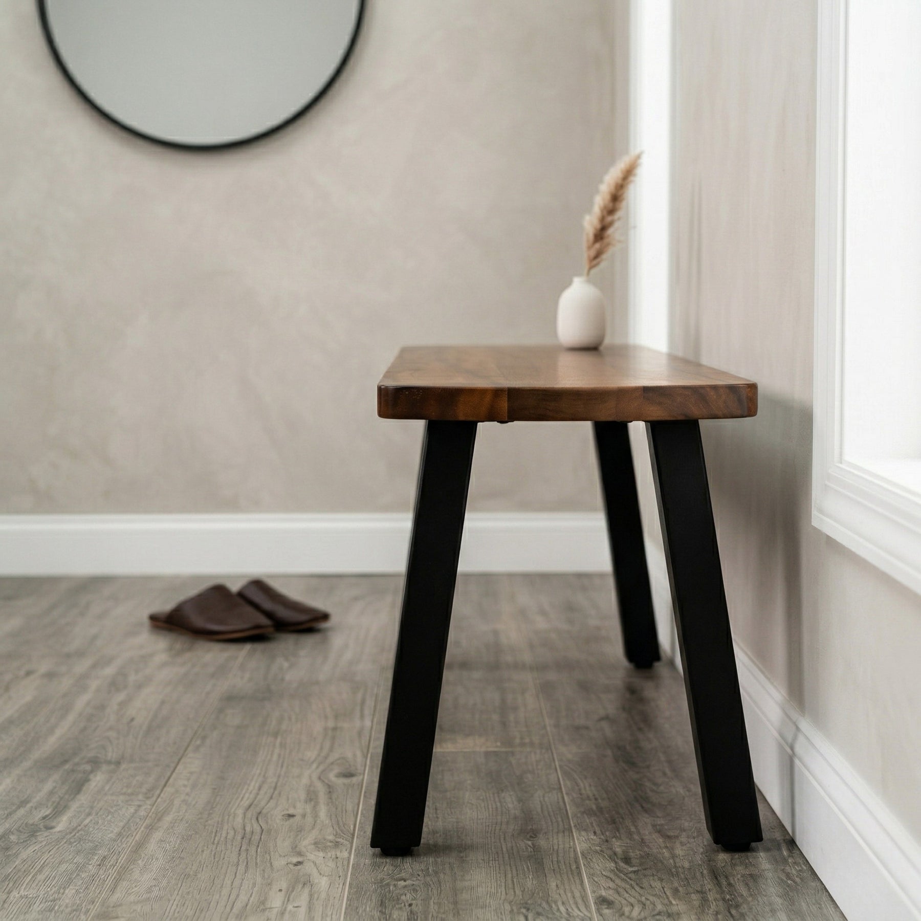 Wooden side bench with black legs in a room with a mirror and vase on a shelf.