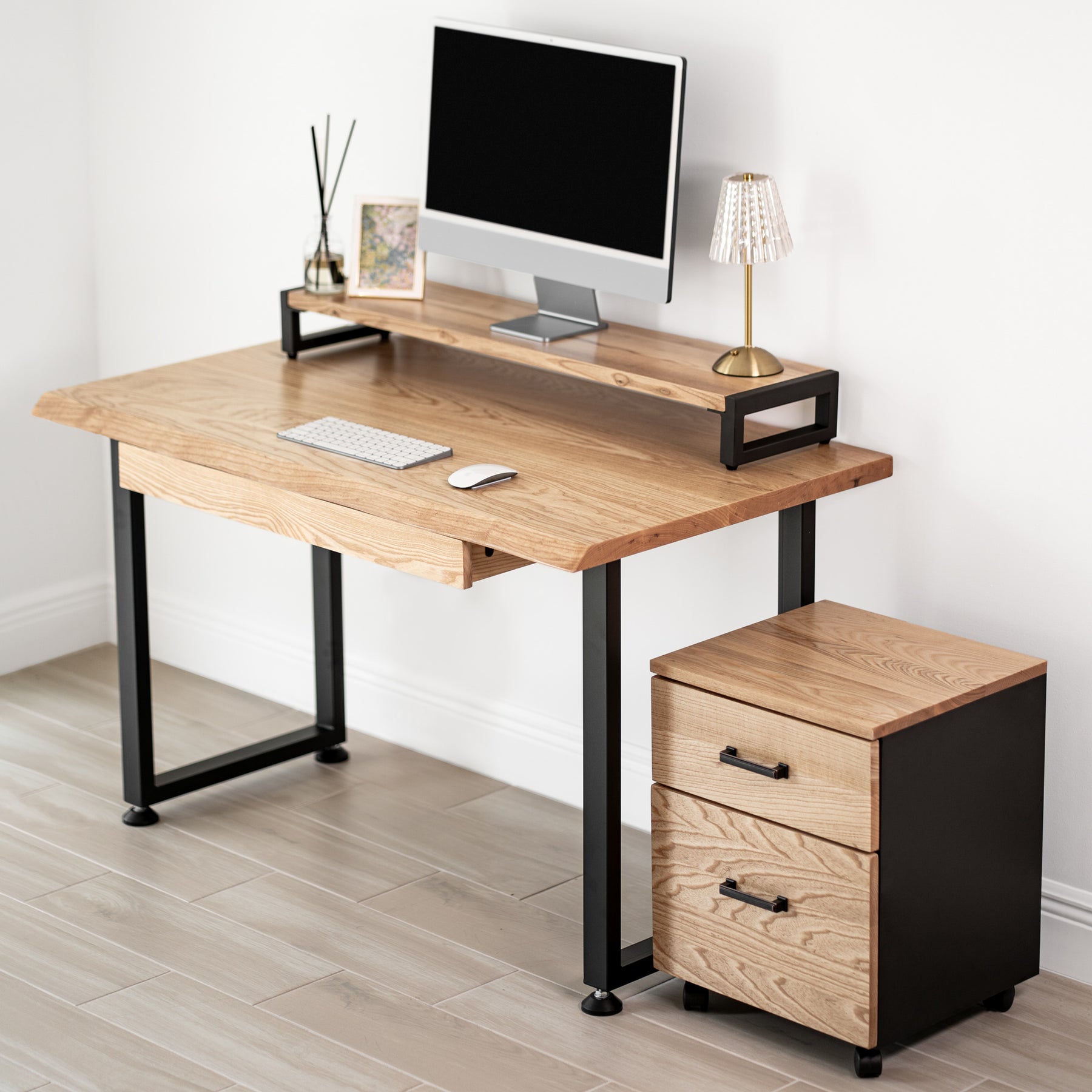 White oak computer desk with a drawer, black metal legs, and a matching storage unit against a white wall.