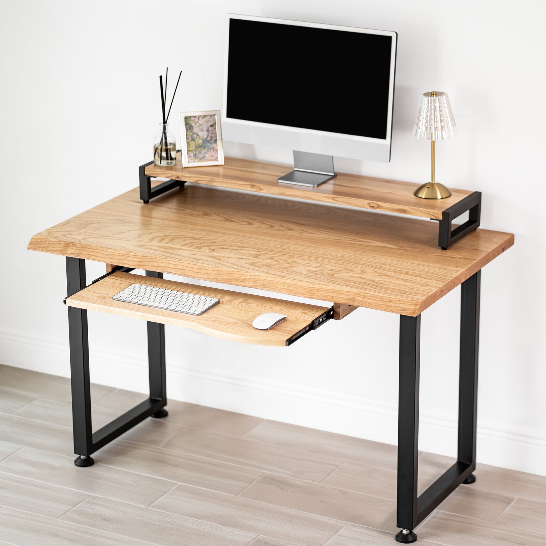 White oak computer desk with keyboard tray, black metal legs, computer monitor stand, keyboard, and lamp on a light wood floor.