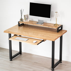White oak computer desk with keyboard tray, black metal legs, computer monitor stand, keyboard, and lamp on a light wood floor.