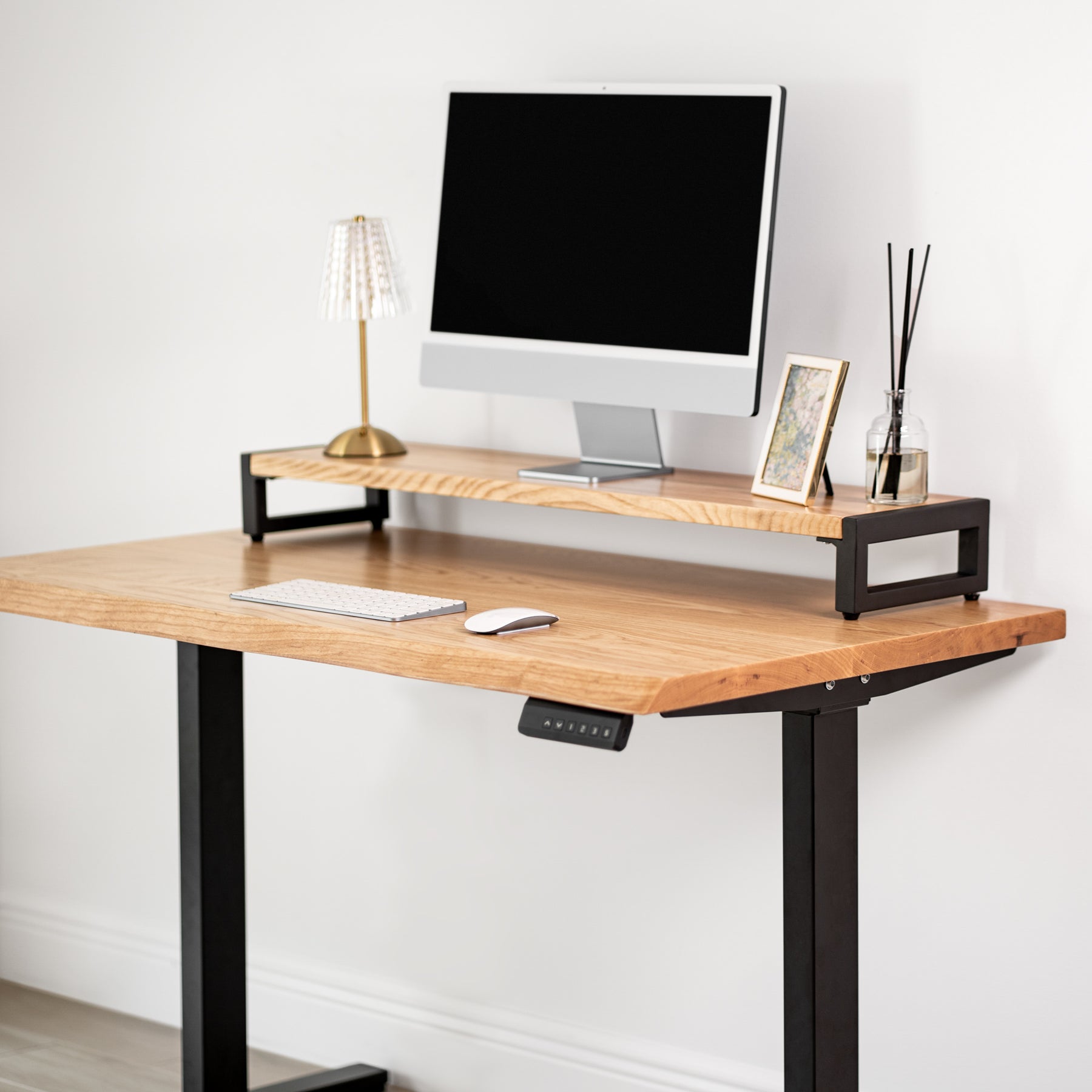 White oak standing desk with black sit-stand legs, computer monitor, keyboard, and decorative items on a white wall background.